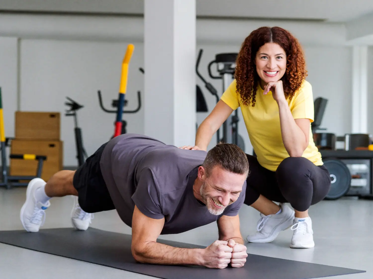 Coach kneeling beside an older man holding a plank on a gym mat